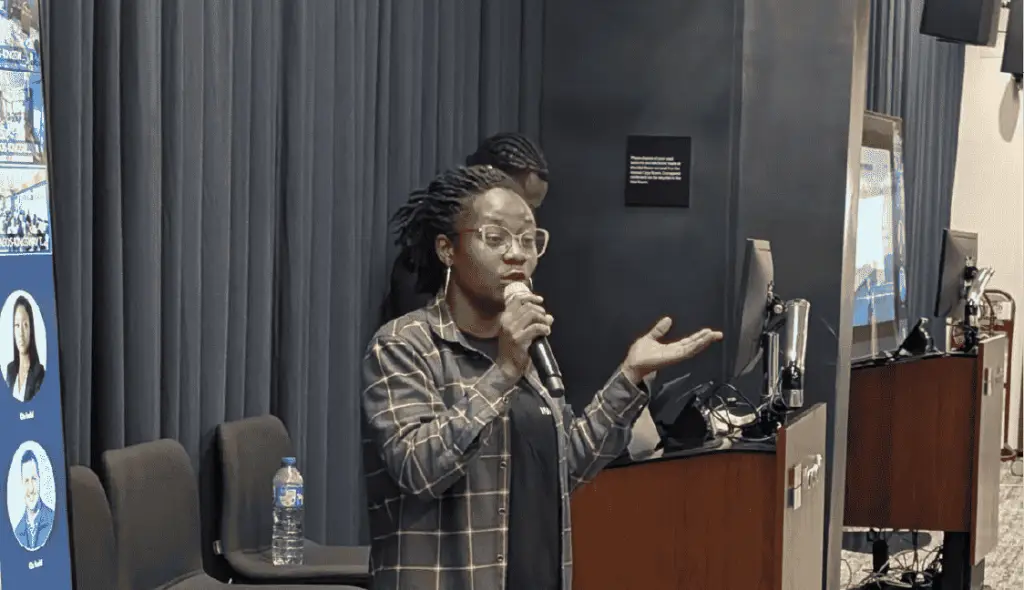 Woman with glasses speaks into a handheld microphone on a stage, gesturing with her other hand.