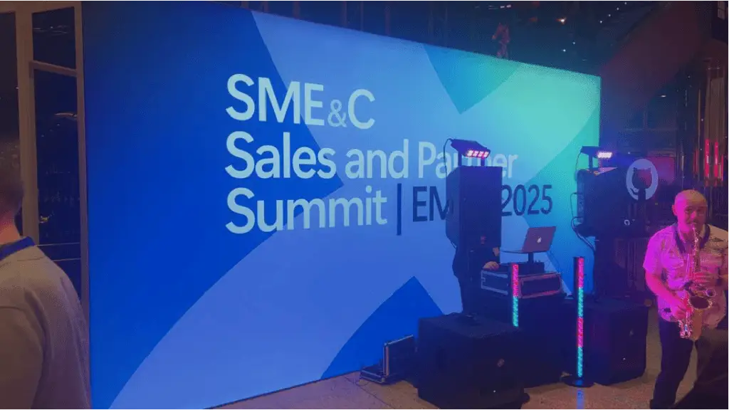 Blue event backdrop reads 'SME&C Sales and Partner Summit' with stage lights and DJ equipment on the left and a saxophonist playing on the right indoors.