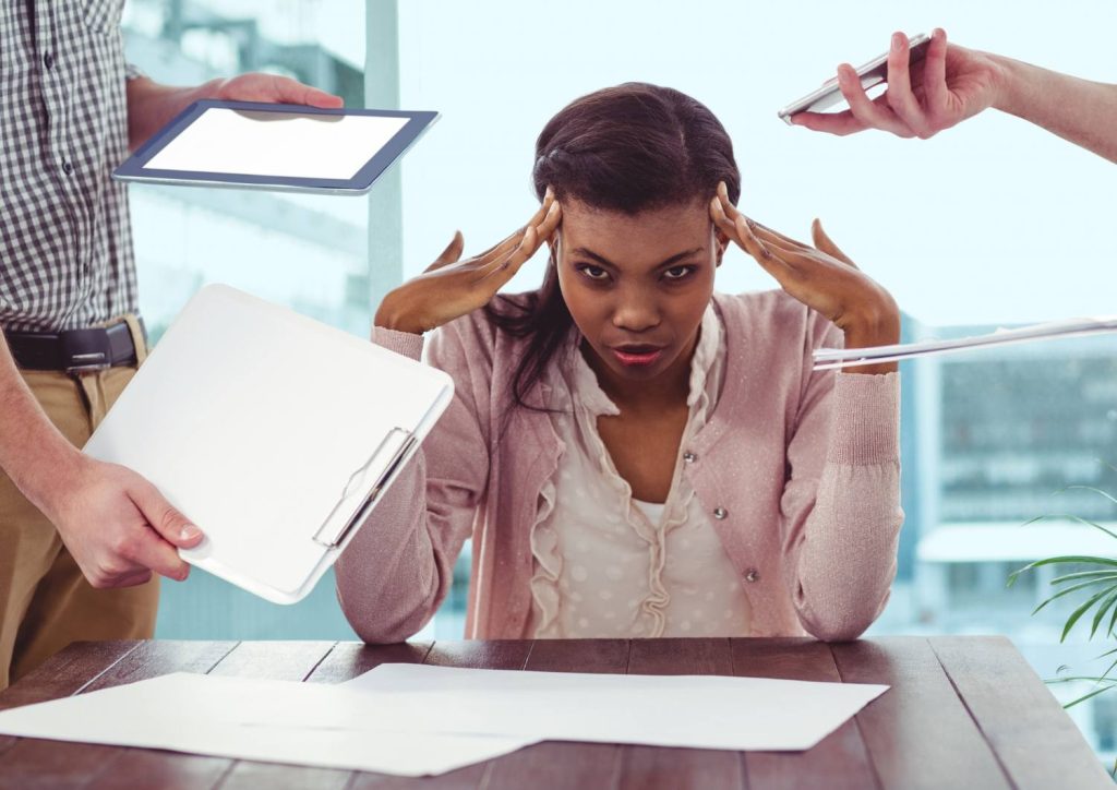 Frustrated woman at a desk during a tense office meeting as teammates hand over a tablet and clipboard with papers on the table.